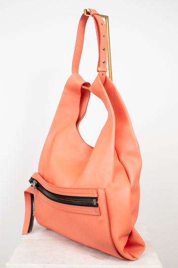 A coral pink leather shoulder bag with a gold-tone metal accent on the strap and a black zipper detail on the front, displayed on a white surface against a white background.