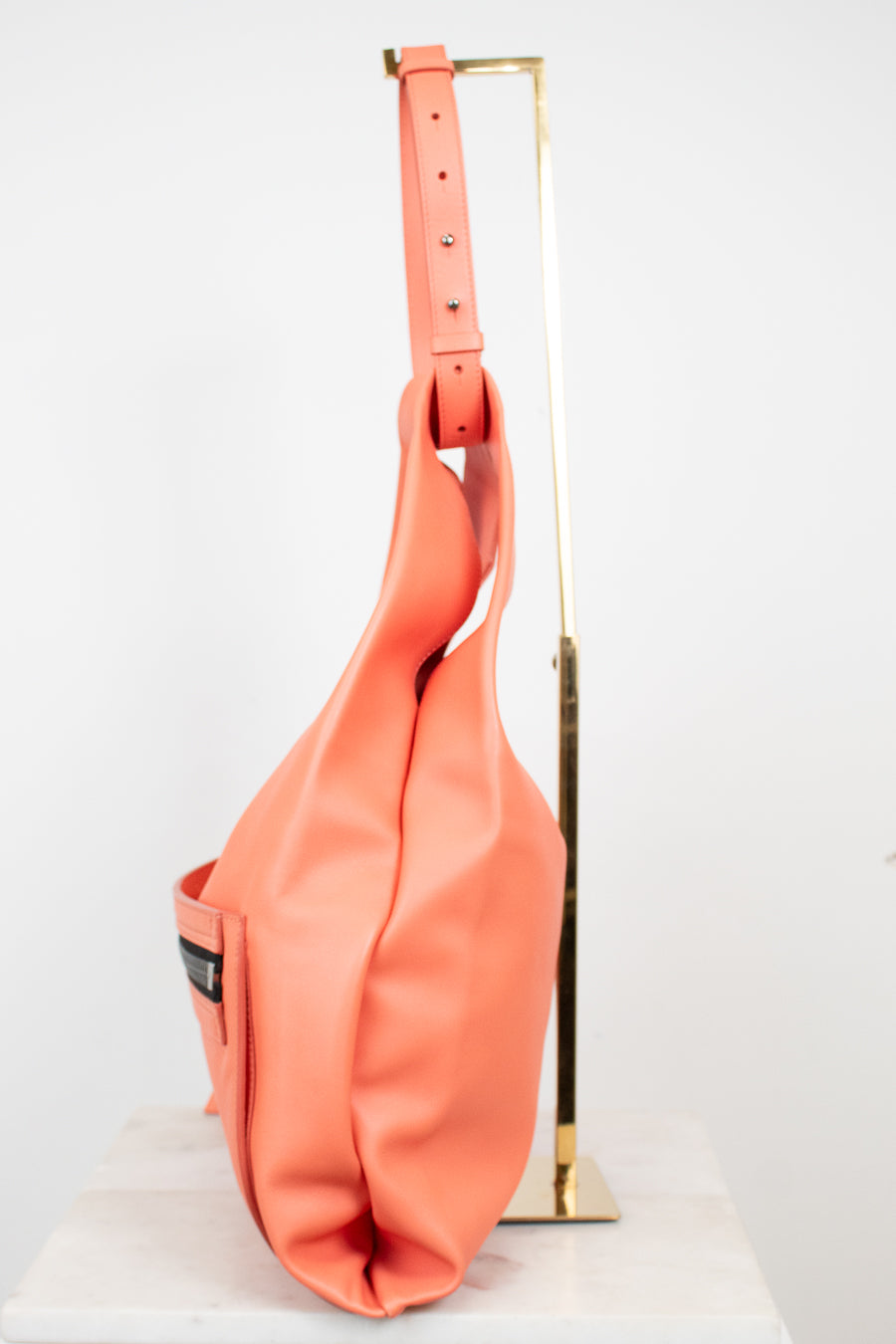 A coral pink leather shoulder bag hanging from a gold metal display stand against a plain white background.