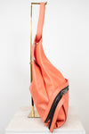 A coral pink leather shoulder bag with black zippers, displayed on a gold metal stand over a white marble block against a white background.