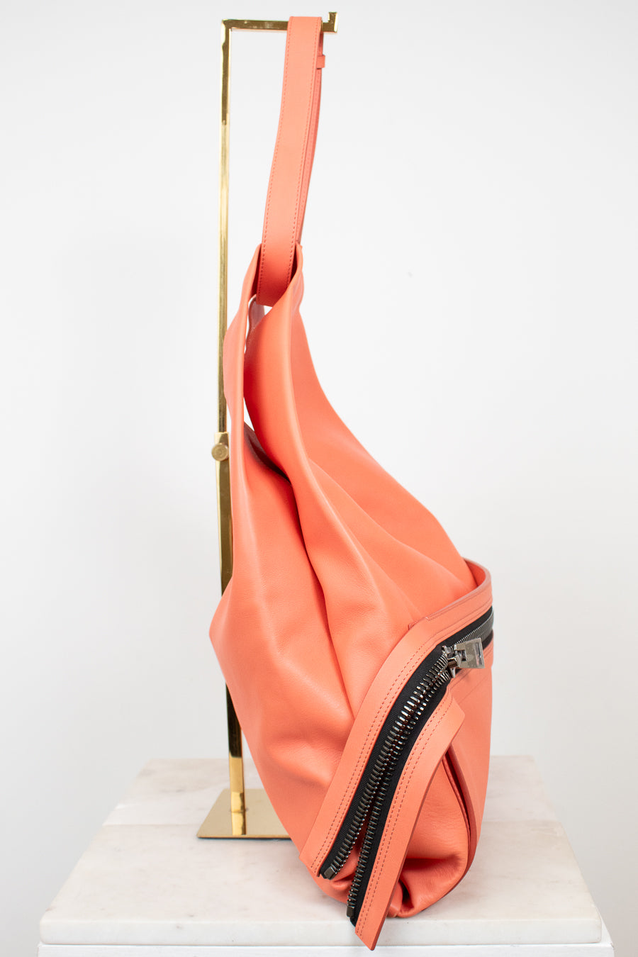 A coral pink leather shoulder bag with black zippers, displayed on a gold metal stand over a white marble block against a white background.