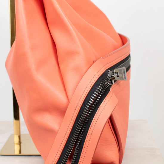 A close-up of a peach-colored faux leather shoulder bag with black double zippers, resting on a white marble surface next to a gold stand.