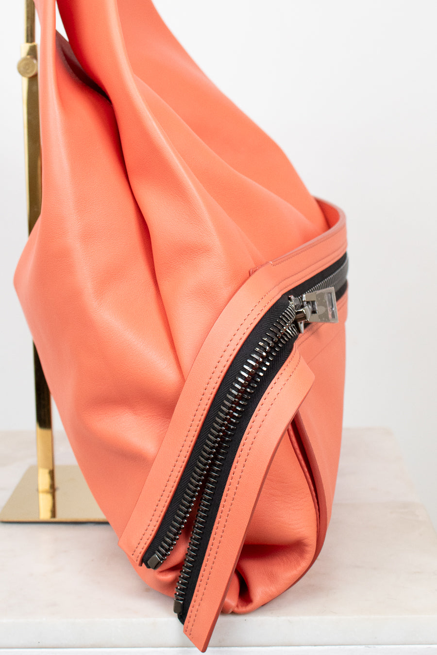 A close-up of a peach-colored faux leather shoulder bag with black double zippers, resting on a white marble surface next to a gold stand.