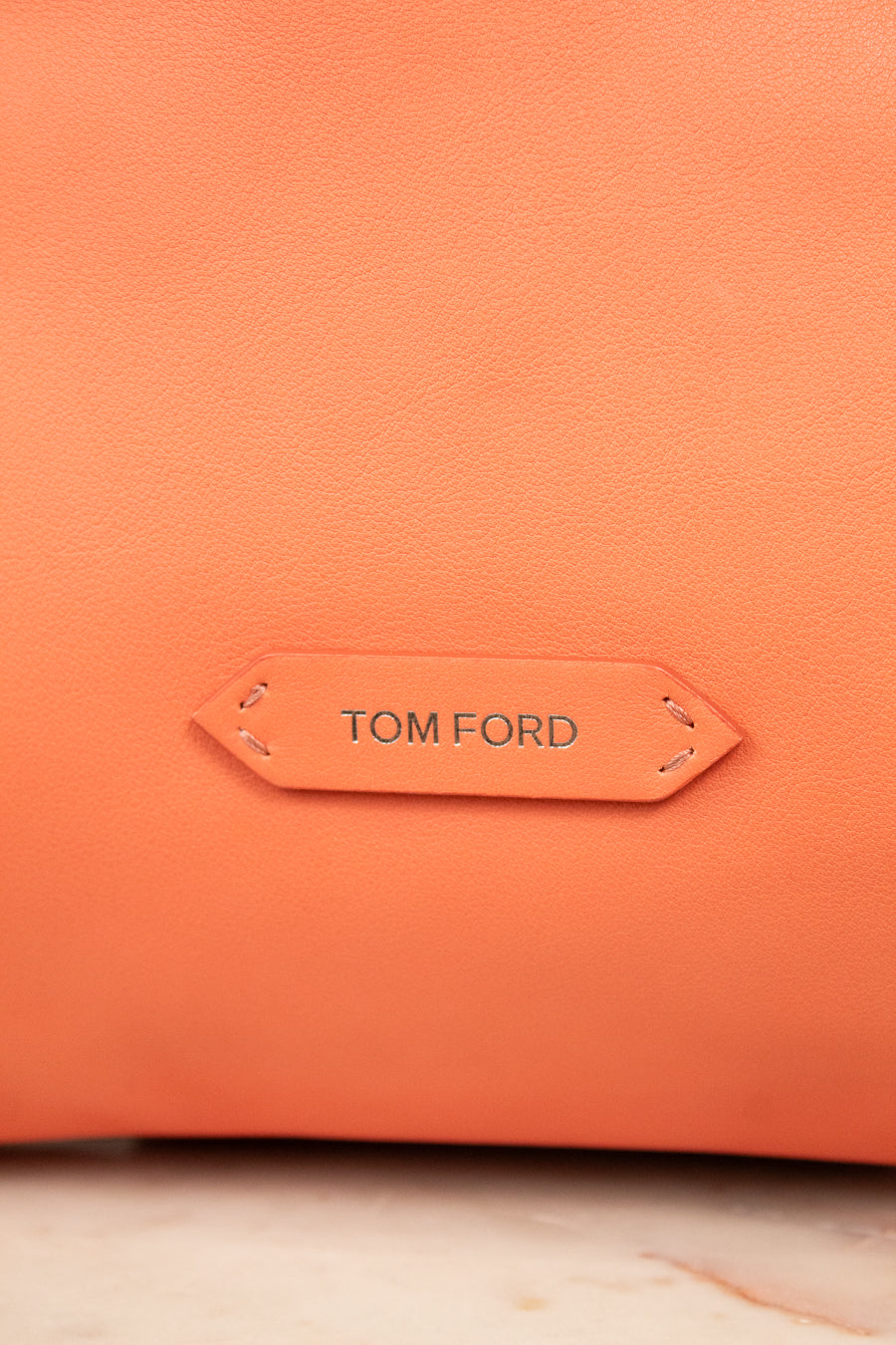 Close-up of a textured orange leather bag with a central tan leather brand tab reading TOM FORD.
