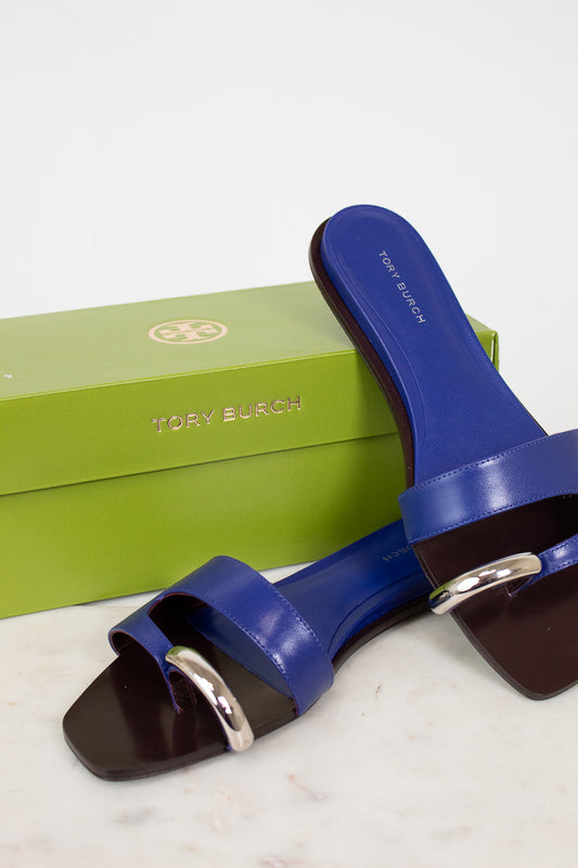 Blue sandals with a silver buckle on a white surface with a green Tory Burch box in the background.