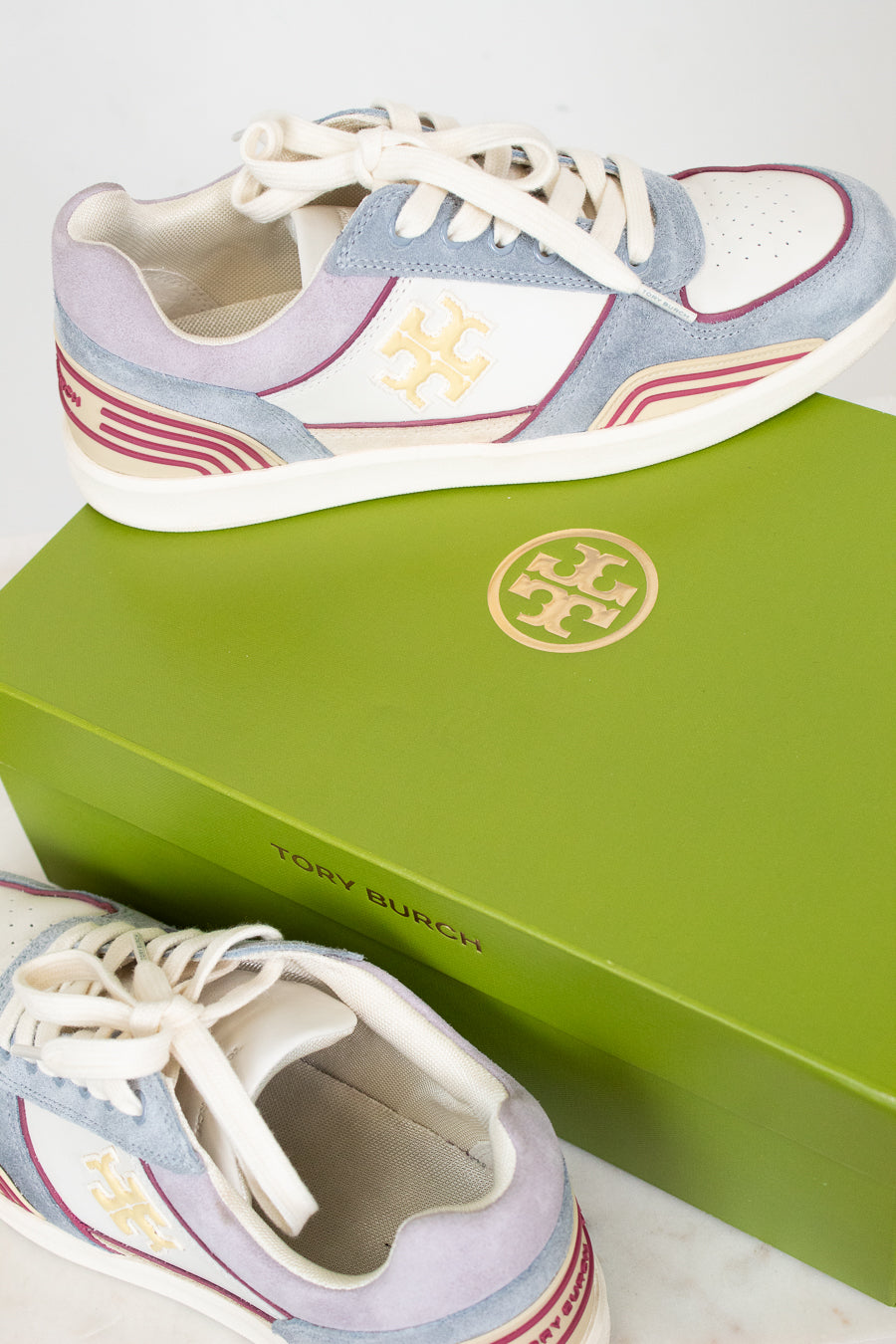 A pair of Tory Burch low-top sneakers in light blue suede and white leather with gold logo details, displayed on top of and next to a bright green branded shoebox.