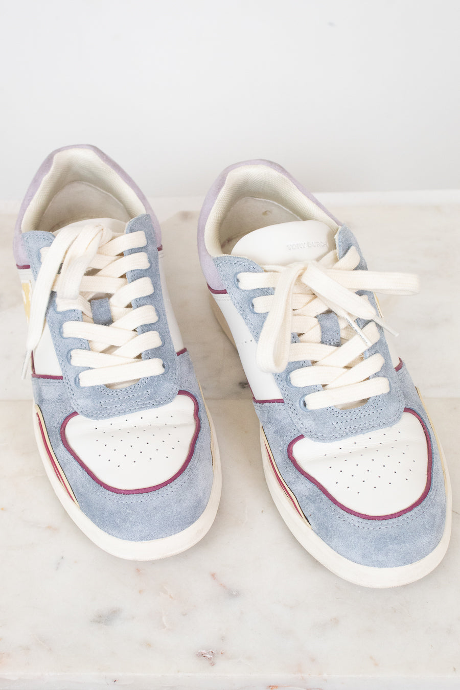 A pair of low-top sneakers in light blue suede and white leather, featuring white laces, a white heart-shaped toe overlay, and thin red piping details, displayed on a white marble surface.