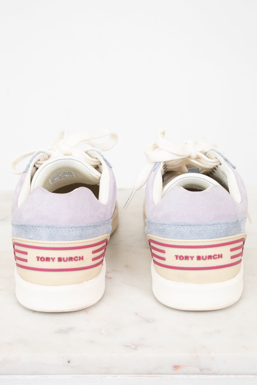 A pair of Tory Burch low-top sneakers shown from the back, featuring lavender suede, light blue suede, cream canvas, and pink stripe accents on the heel.