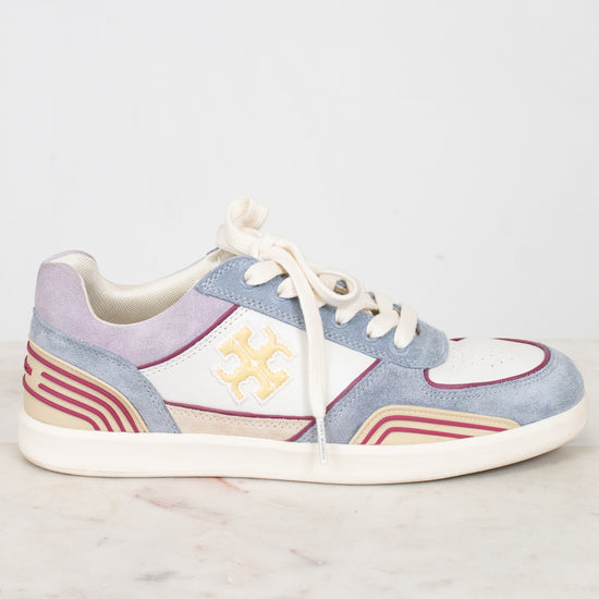 A low-top sneaker with a light blue suede upper, cream leather panels, cream laces, and a cream sole, featuring a gold monogram logo on the side. The shoe is displayed on a light marble surface against a white background.