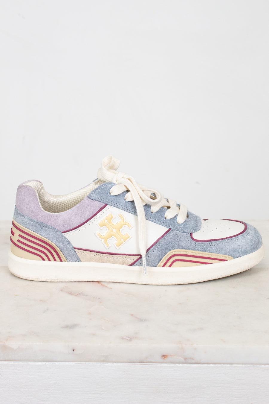 A low-top sneaker with a light blue suede upper, cream leather panels, cream laces, and a cream sole, featuring a gold monogram logo on the side. The shoe is displayed on a light marble surface against a white background.