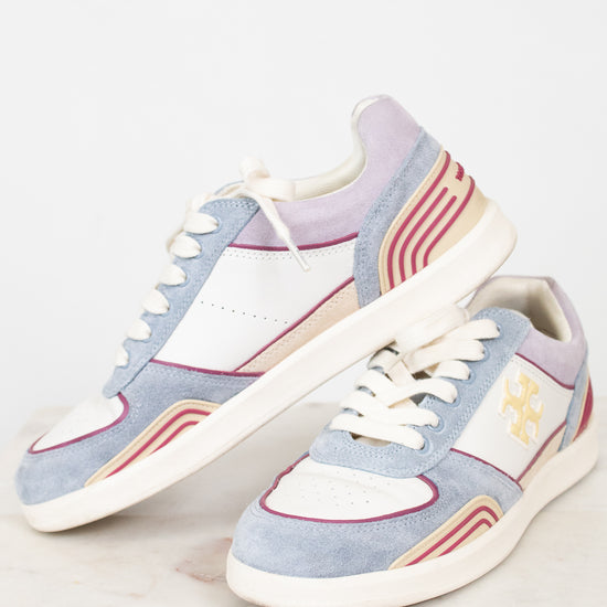 A pair of low-top sneakers in a pastel colorway featuring light blue suede, white leather panels, lavender purple overlays, and beige and red wavy stripe accents. One shoe is positioned upright and the other lies on its side, showcasing the side profile with a gold monogram logo on a clean, light marble surface against a white background.