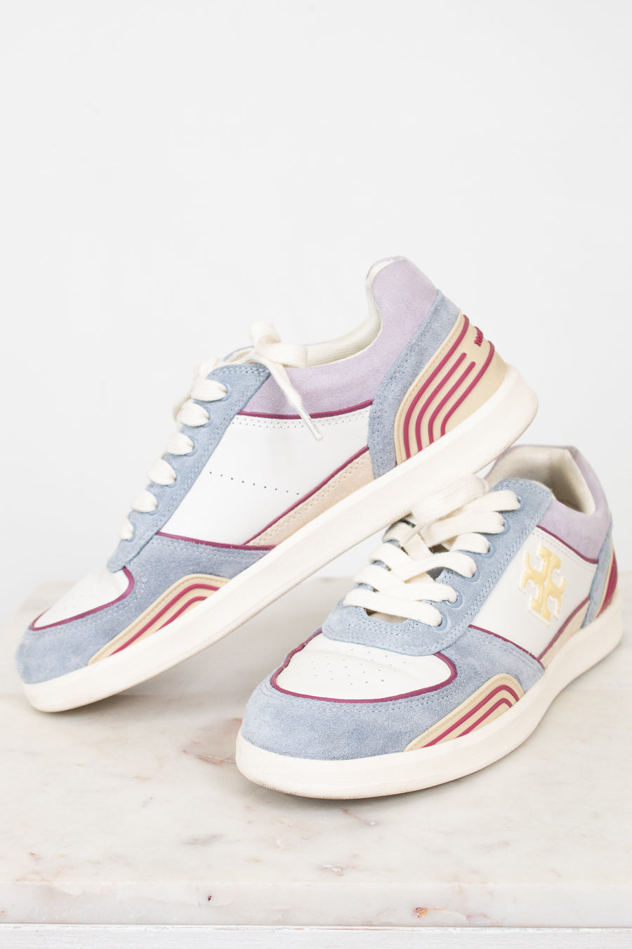 A pair of low-top sneakers in a pastel colorway featuring light blue suede, white leather panels, lavender purple overlays, and beige and red wavy stripe accents. One shoe is positioned upright and the other lies on its side, showcasing the side profile with a gold monogram logo on a clean, light marble surface against a white background.