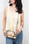 A woman wearing a pale yellow sleeveless tie-front blouse and blue jeans, holding a snakeskin-patterned shoulder bag with a gold-tone logo clasp against a plain light background.