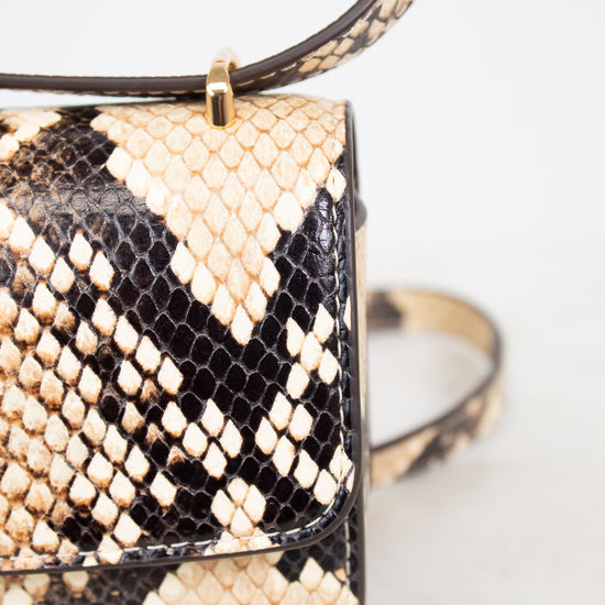 Close-up of a snakeskin leather handbag with a flap front, gold-tone hardware, and a strap, photographed against a light neutral background.