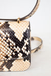 Close-up of a snakeskin leather handbag with a flap front, gold-tone hardware, and a strap, photographed against a light neutral background.