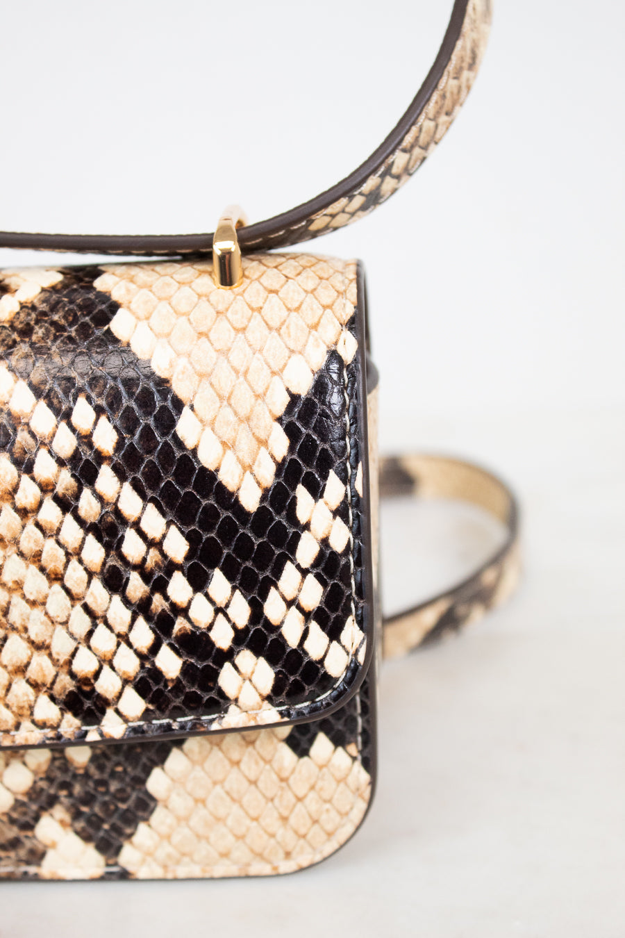 Close-up of a snakeskin leather handbag with a flap front, gold-tone hardware, and a strap, photographed against a light neutral background.