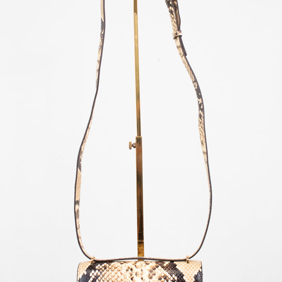 A small snakeskin-pattern shoulder bag with a gold-tone logo clasp and a long matching strap, displayed on a gold stand against a white background.