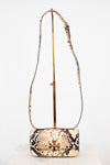 A small snakeskin-pattern shoulder bag with a gold-tone logo clasp and a long matching strap, displayed on a gold stand against a white background.