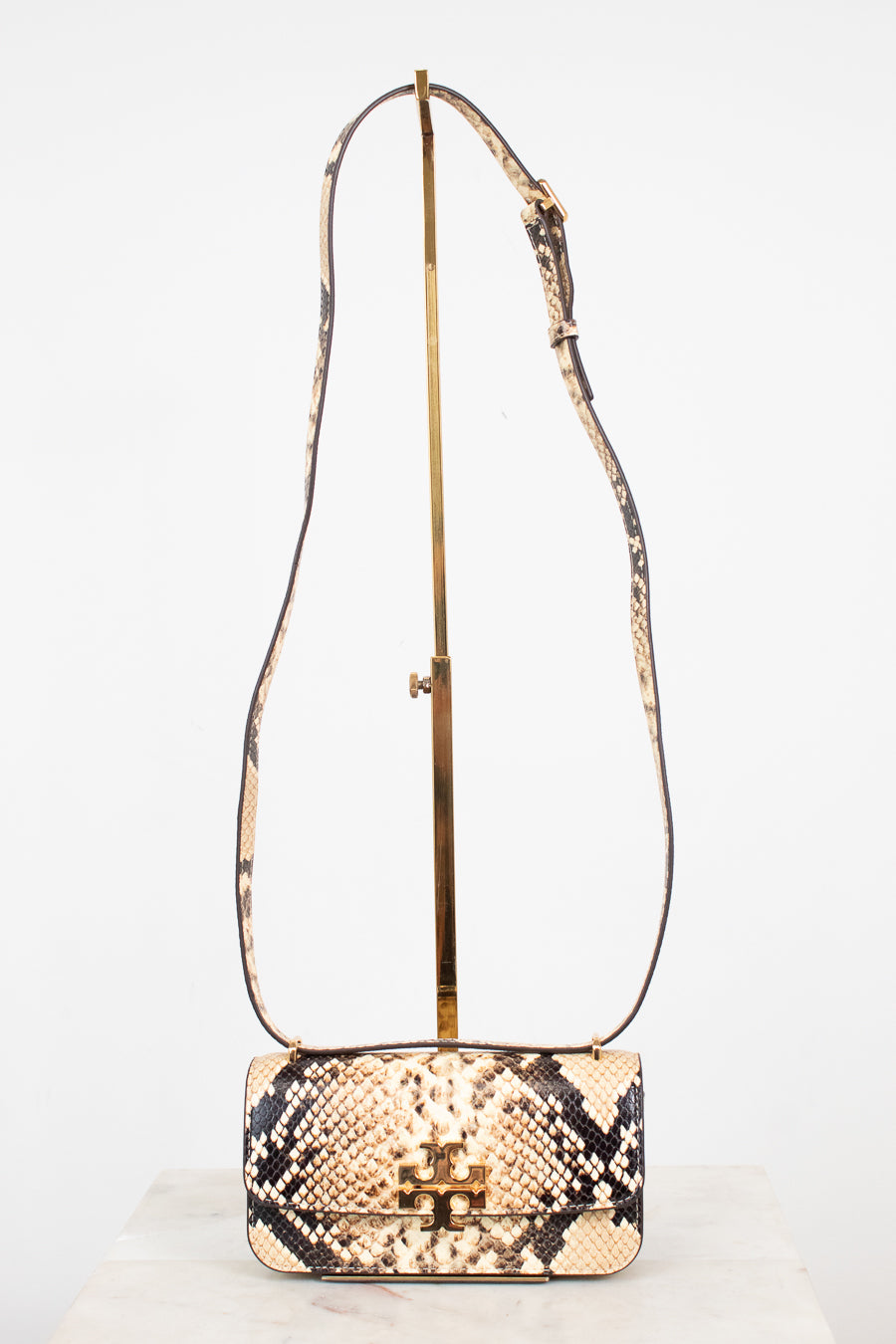 A small snakeskin-pattern shoulder bag with a gold-tone logo clasp and a long matching strap, displayed on a gold stand against a white background.
