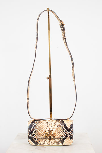 A small snakeskin-pattern shoulder bag with a gold-tone logo clasp and a long matching strap, displayed on a gold stand against a white background.