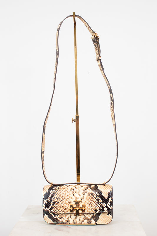 A small snakeskin-pattern shoulder bag with a gold-tone logo clasp and a long matching strap, displayed on a gold stand against a white background.