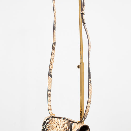 A snakeskin leather shoulder bag with a long matching strap on a gold-tone metal stand, displayed on a white marble surface against a white background.
