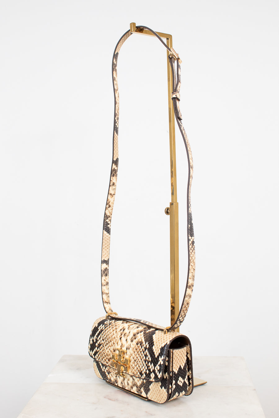 A snakeskin leather shoulder bag with a long matching strap on a gold-tone metal stand, displayed on a white marble surface against a white background.