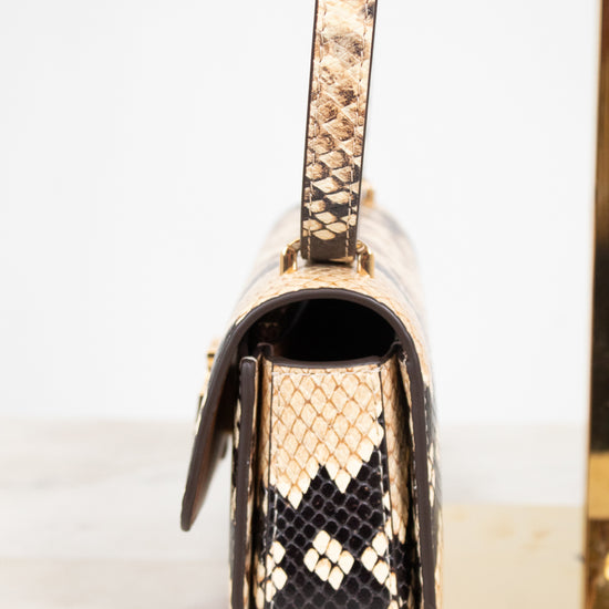 A close-up side view of a snakeskin leather handbag,  featuring a gold-tone metal frame and strap attachment, set against a clean white background.
