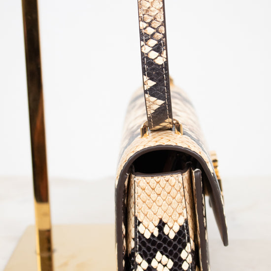A close-up side view of a snakeskin leather handbag with a flap closure and gold-tone hardware, displayed on a gold stand against a white background.