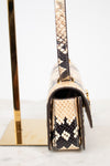 A close-up side view of a snakeskin leather handbag with a flap closure and gold-tone hardware, displayed on a gold stand against a white background.