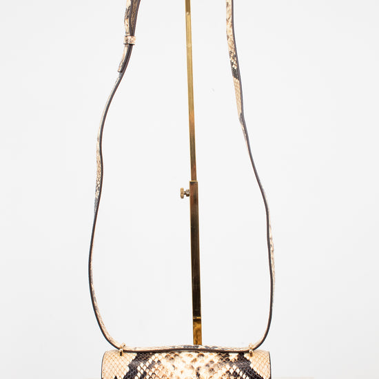 A snakeskin leather shoulder bag with a rectangular body and long matching  strap, displayed on a gold stand against a white background.