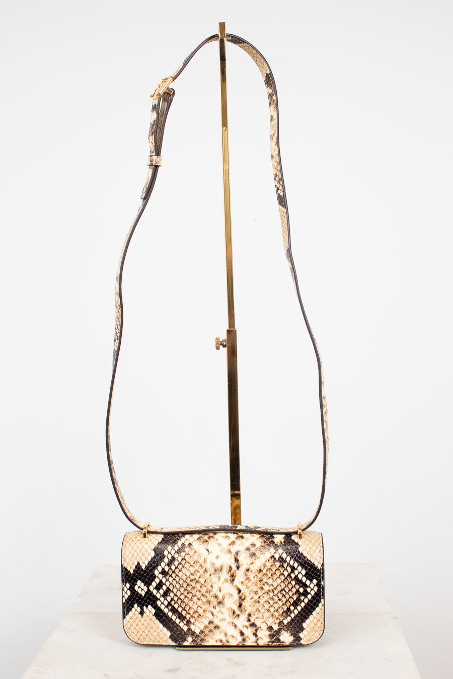 A snakeskin leather shoulder bag with a rectangular body and long matching  strap, displayed on a gold stand against a white background.