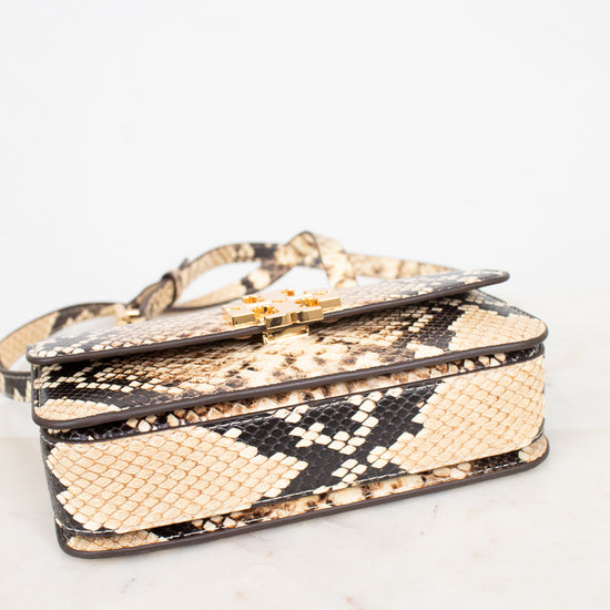 A snakeskin leather handbag with a flap closure and gold-tone hardware, resting on a light marble surface against a white background.