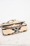 A snakeskin leather handbag with a flap closure and gold-tone hardware, resting on a light marble surface against a white background.