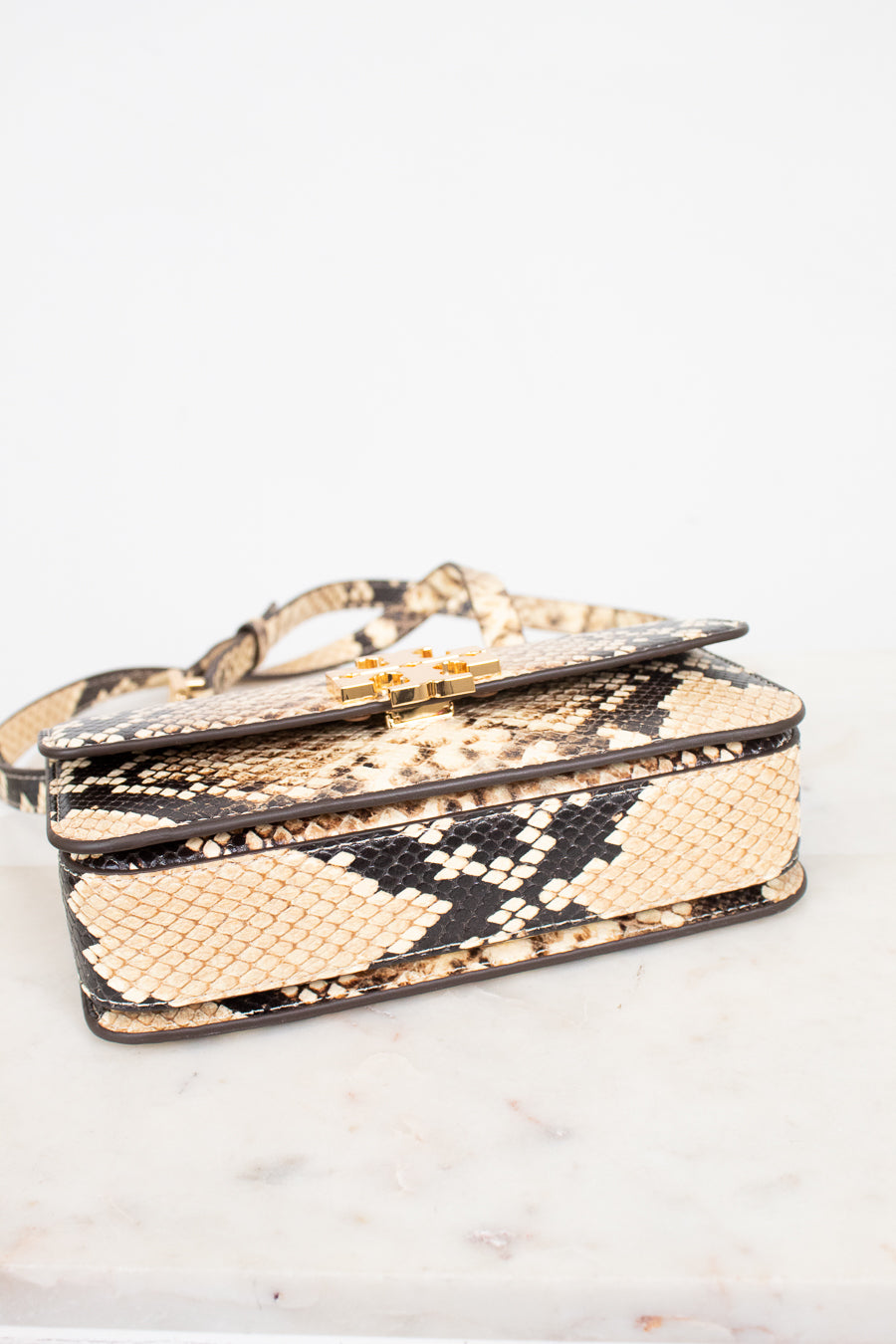 A snakeskin leather handbag with a flap closure and gold-tone hardware, resting on a light marble surface against a white background.