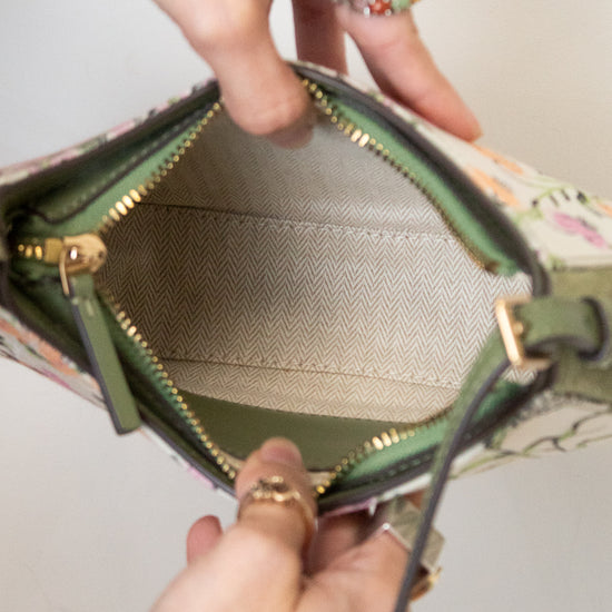 Close-up of a handbag being opened by two hands, showing the interior lining, green leather trim, gold zipper, and a floral patterned exterior against a plain light background.