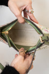 Close-up of a handbag being opened by two hands, showing the interior lining, green leather trim, gold zipper, and a floral patterned exterior against a plain light background.