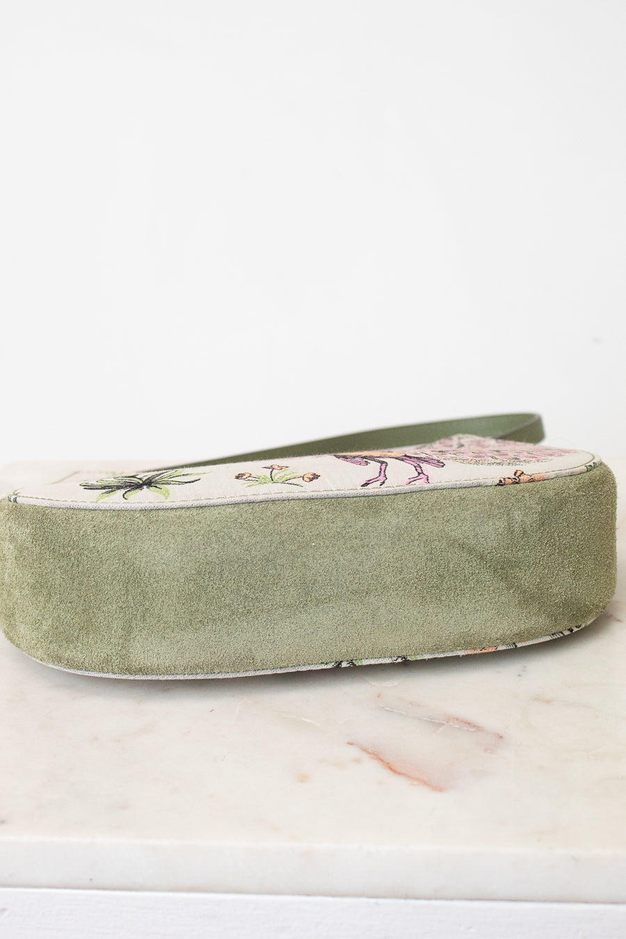 An olive green velvet shoe bag with a floral-patterned fabric top panel, resting on a white marble surface against a white background.