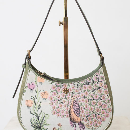 A small crescent-shaped shoulder bag with a peacock and floral print on a light background, displayed on a gold-colored stand against a white backdrop.