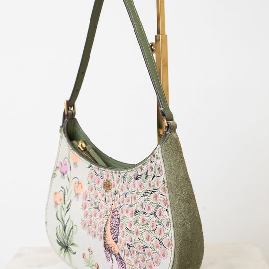 An olive green hobo shoulder bag with a detailed peacock and floral print on the front, displayed on a gold metal stand against a white background.