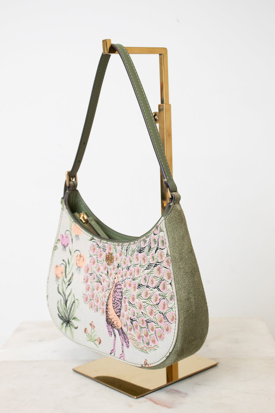 An olive green hobo shoulder bag with a detailed peacock and floral print on the front, displayed on a gold metal stand against a white background.