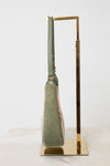 A structured shoulder bag in sage green suede with a light pink patterned lining, displayed on a gold metal stand against a white background.