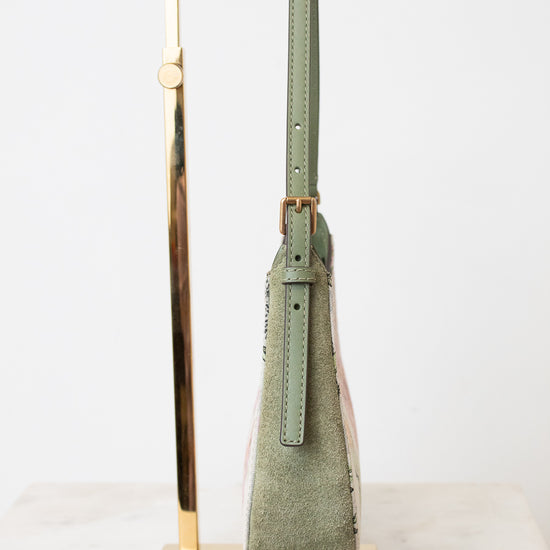 A sage green suede shoulder bag with a matching leather strap and gold-tone hardware, displayed on a gold metal stand against a white background.