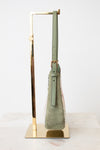A sage green suede shoulder bag with a matching leather strap and gold-tone hardware, displayed on a gold metal stand against a white background.