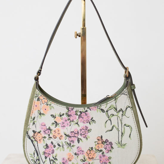 A crescent-shaped shoulder bag with a white base, featuring a colorful floral and botanical print in pink, purple, orange, and green. The bag has dark brown leather trim and a single shoulder strap, displayed on a gold-colored metal stand against a plain white background.