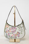 A crescent-shaped shoulder bag with a white base, featuring a colorful floral and botanical print in pink, purple, orange, and green. The bag has dark brown leather trim and a single shoulder strap, displayed on a gold-colored metal stand against a plain white background.