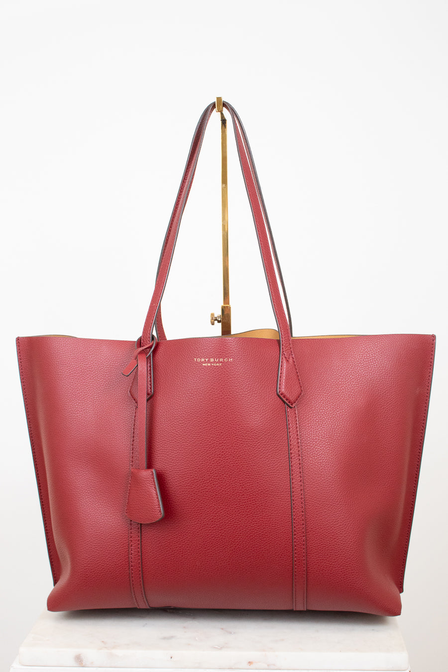 A red leather tote bag with double shoulder straps and a hanging leather tag, displayed on a gold stand against a white background.