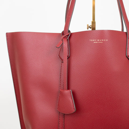 A red pebbled leather tote bag by Tory Burch, featuring dual shoulder straps, a hanging leather tag, and gold-tone hardware, shown against a clean white background.
