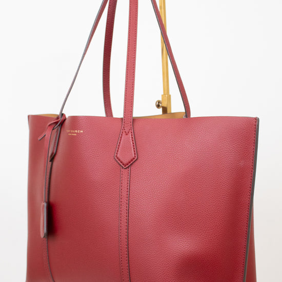 A red pebbled leather tote bag with double shoulder straps, displayed on a gold metal stand against a clean white background.