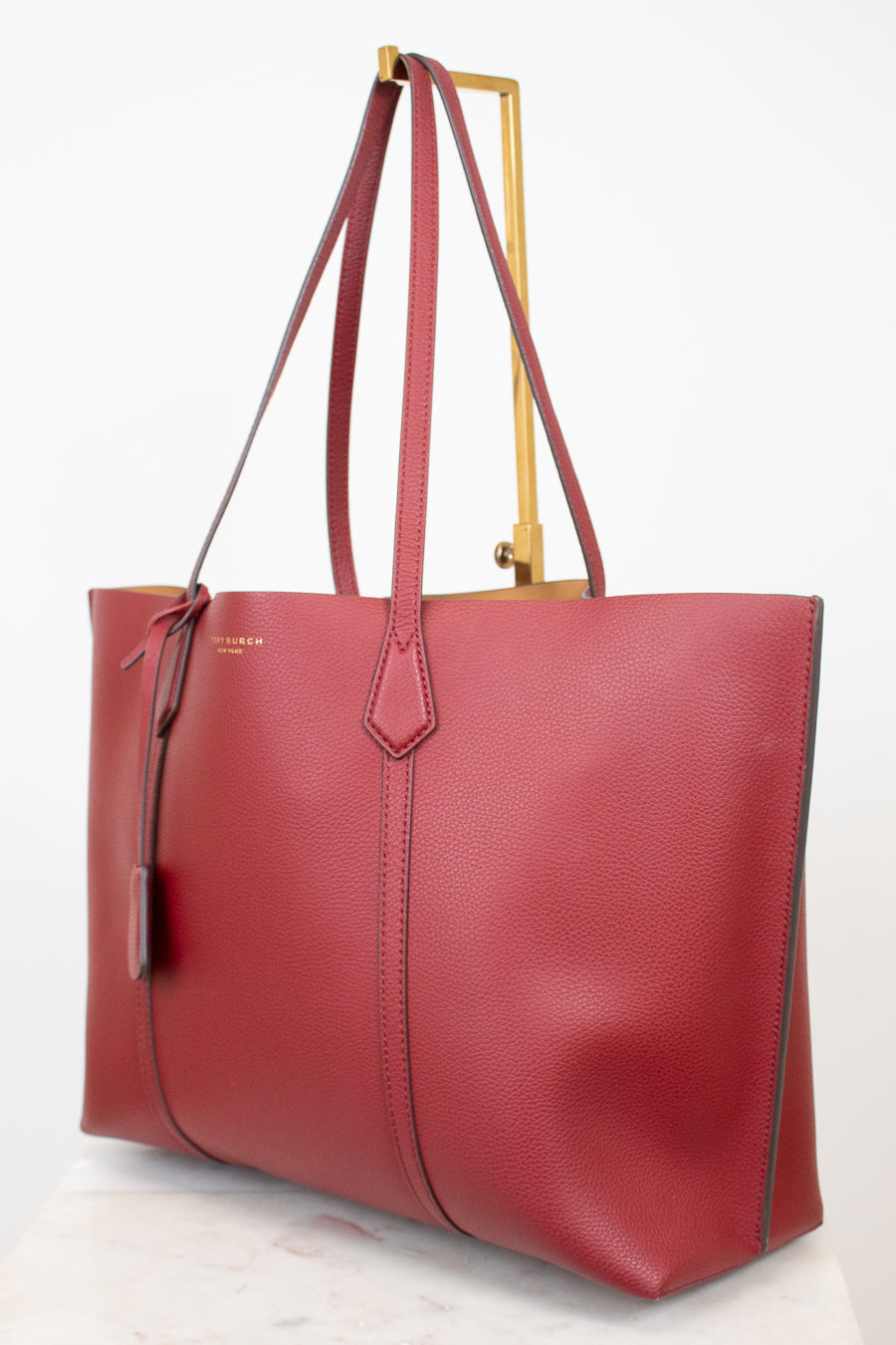 A red pebbled leather tote bag with double shoulder straps, displayed on a gold metal stand against a clean white background.