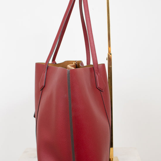A red leather tote bag with a pebbled texture, displayed on a white marble surface next to a gold metal stand against a white background.